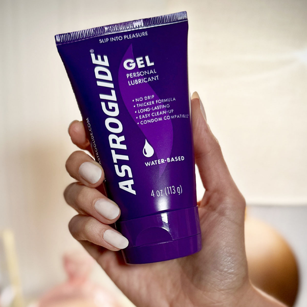 A close-up, stylized photo of a hand holding the purple tube of Astroglide Gel Personal Lubricant in an indoor setting.