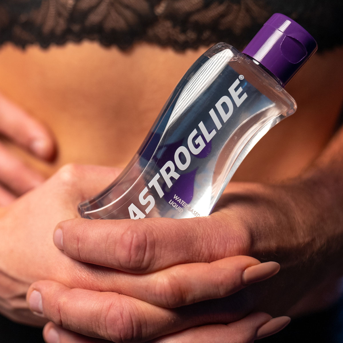 A close-up of a clear bottle of Astroglide Water-Based Liquid personal lubricant, held in two hands against a blurred background of a person wearing black lace lingerie.