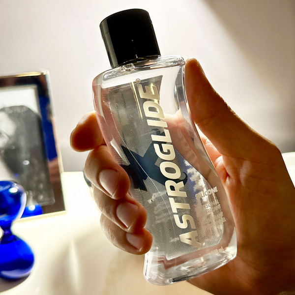 A hand holding the clear bottle of Astroglide X Premium Silicone Liquid, with a blurred blue hourglass in the background.