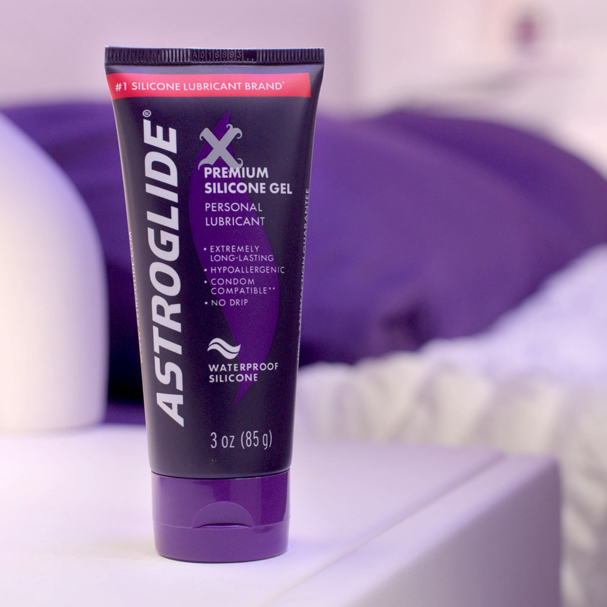 A tube of Astroglide X Premium Silicone Gel on a white surface next to a bed with purple bedding.