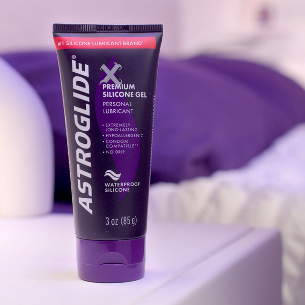 A tube of Astroglide X Premium Silicone Gel on a white surface next to a bed with purple bedding.