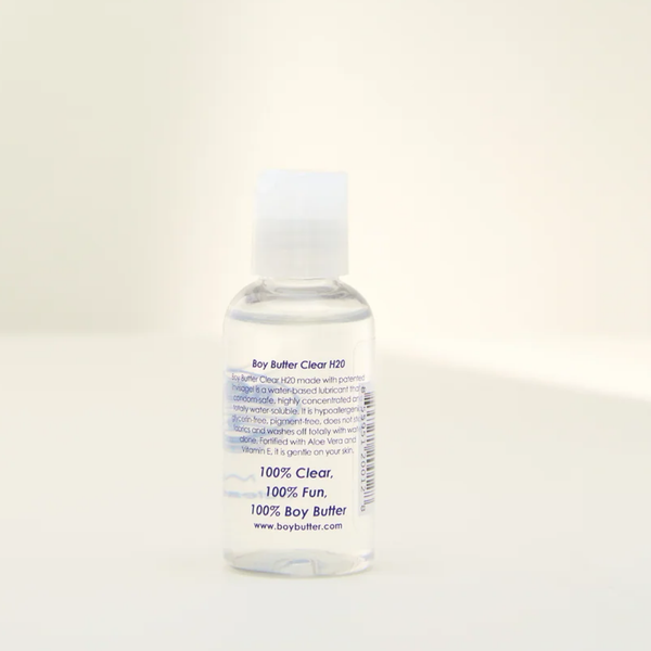 Side view of the Boy Butter CLEAR lubricant bottle, showing a clear liquid inside. Text on the label describes the product as 'Boy Butter Clear H2O' a water-based lubricant, 'condom-safe,' 'hypoallergenic,' and '100% Clear, 100% Fun, 100% Boy Butter'