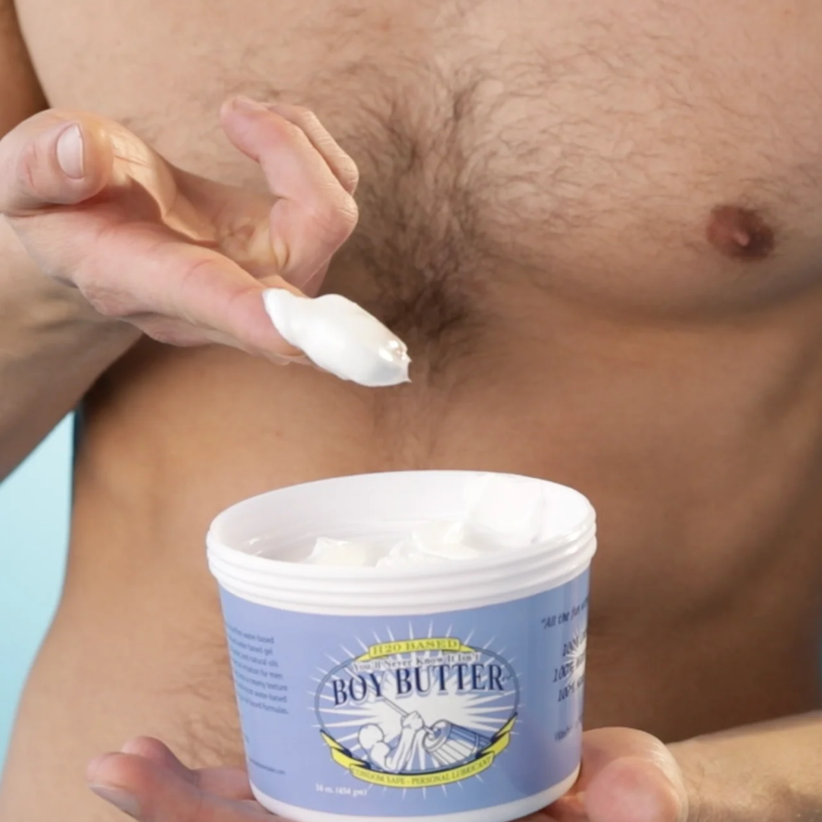 A close-up shot of a man's bare torso and hand, showing a white dollop of Boy Butter H2O Based lubricant on a fingertip being pulled from an open, blue-labeled 16 oz tub.