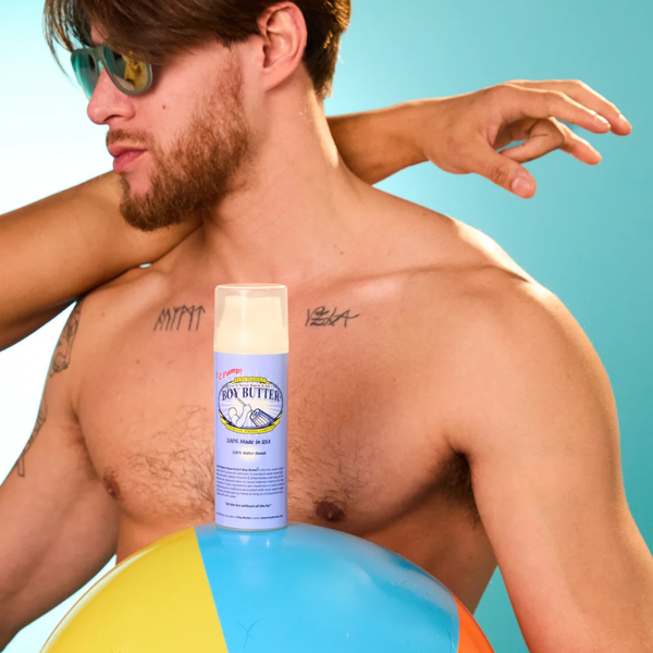 A close-up shot of a shirtless man wearing sunglasses against a bright blue background, with another man's arm around his neck. A small pump bottle of Boy Butter H2O Based lubricant is resting on a colorful beach ball in the foreground.