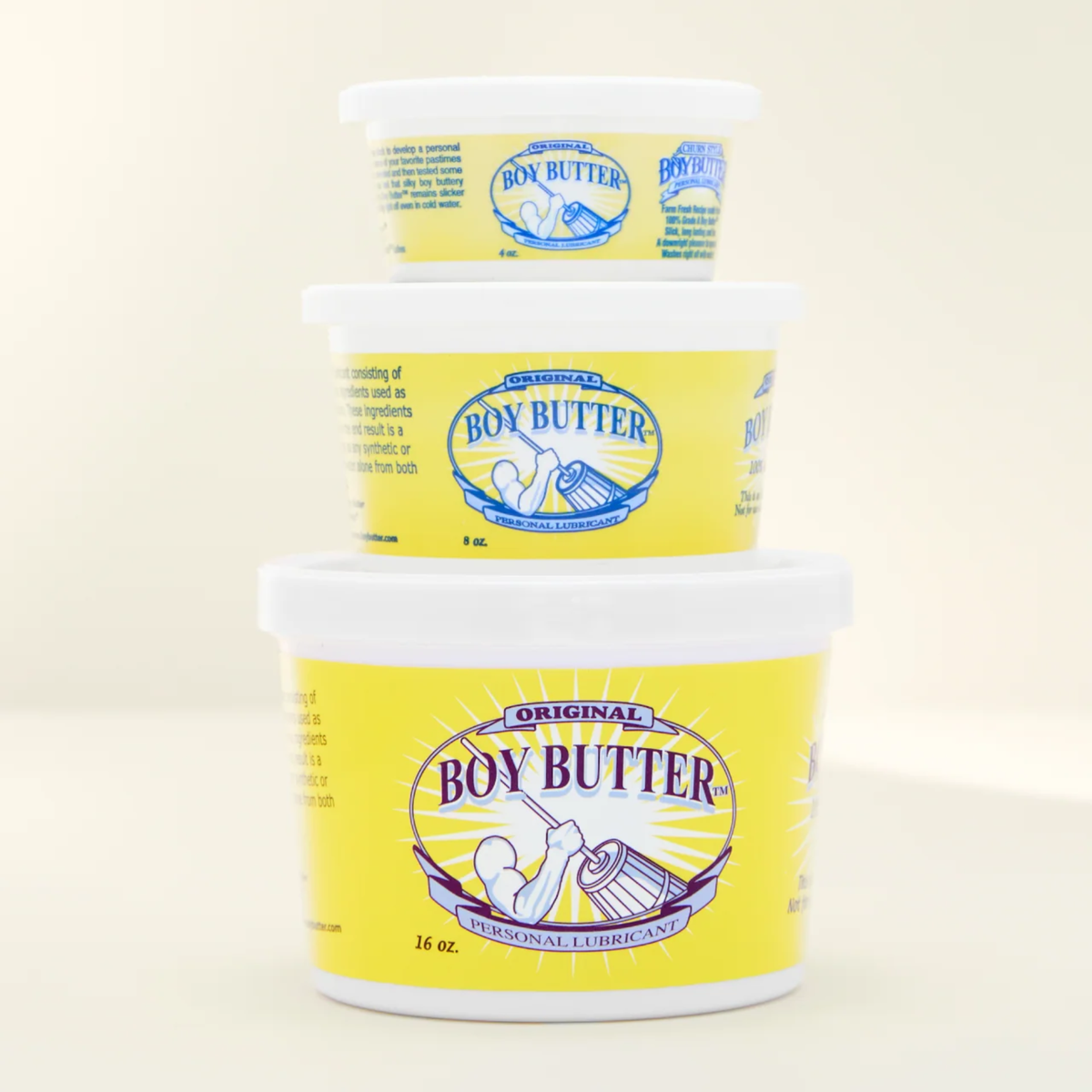 A shot showing a stack of three white plastic tubs of Boy Butter Original Personal Lubricant against a light cream-colored background. The tubs are stacked largest (16 oz.) on the bottom, medium (8 oz.) in the middle, and smallest (2 oz.) on top, each featuring the yellow label with the image of a person churning butter.