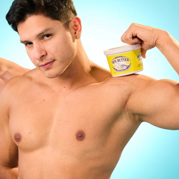 A close-up shot of a muscular man against a bright blue background, looking directly at the camera with a slight smirk. He is shirtless and has a small gold earring. He is holding a yellow tub of Boy Butter Original lubricant on his right shoulder.