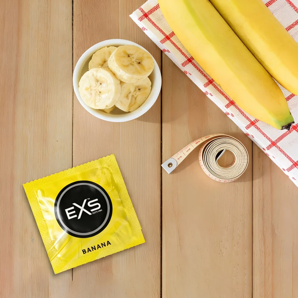 Flat lay photo on a wooden table featuring a single yellow EXS Banana condom wrapper next to fresh whole bananas, sliced banana pieces in a small white bowl, and a measuring tape.