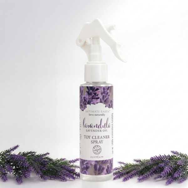 Nature-inspired studio shot of the Intimate Earth Lavandula lavender oil toy cleaner spray bottle flanked by artificial lavender sprigs.
