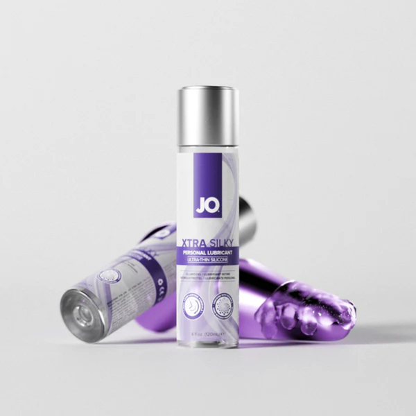 A studio product shot showing a bottle of JO Xtra Silky Personal Lubricant standing upright on a white surface. An additional bottle and toy lie horizontally in the blurred background.
