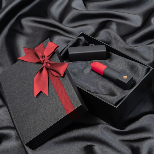 Gift presentation shot showing the red-tipped Lovense Exomoon lipstick vibrator and its black cap lying inside a silk-lined black gift box tied with a red ribbon.