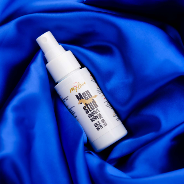 The 50 ml bottle of MyLove Men Stud Comfort Delay Med Spray rests diagonally on draped, rich royal blue satin fabric.