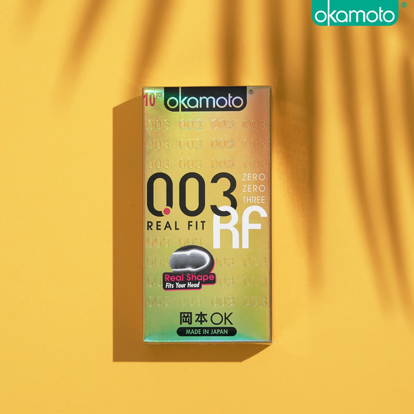 A top-down photo of the shimmering gold-toned Okamoto 003 Real Fit box on a yellow background with palm leaf shadows. A pink sticker on the box says "Real Shape Fits Your Head."