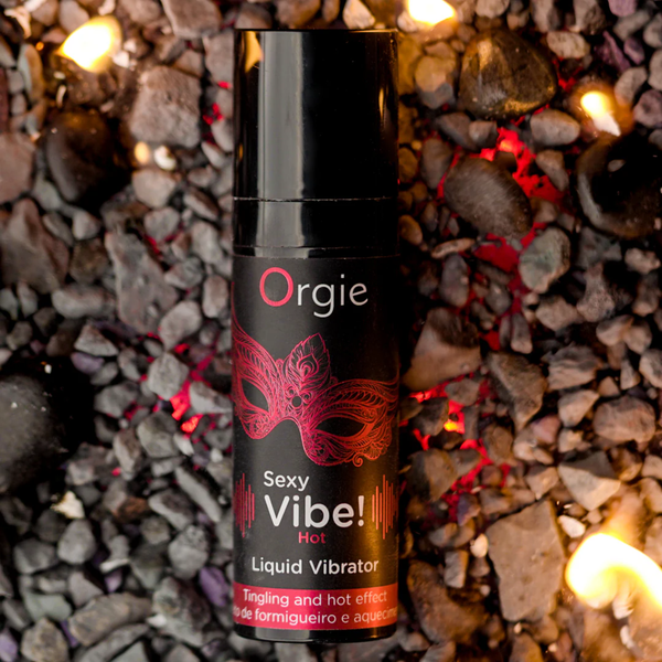 A close-up of the small black pump bottle of Orgie Sexy Vibe! Hot Liquid Vibrator with a red mask design, placed on dark grey rocks with glowing embers around it, symbolizing the hot effect.