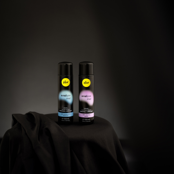 Two bottles of pjur "analyse me!" lubricant are positioned on a draped black cloth against a dark background. 