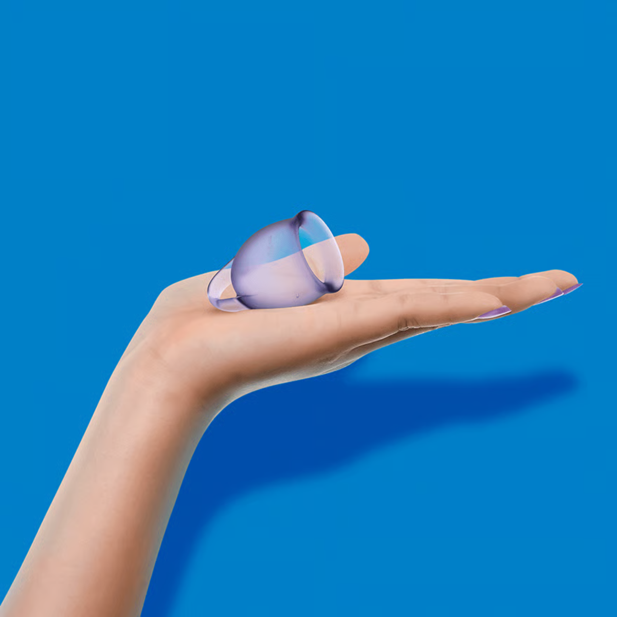 Scale perspective photo showing a translucent lilac silicone menstrual cup held upright on an outstretched hand against a bright blue background.