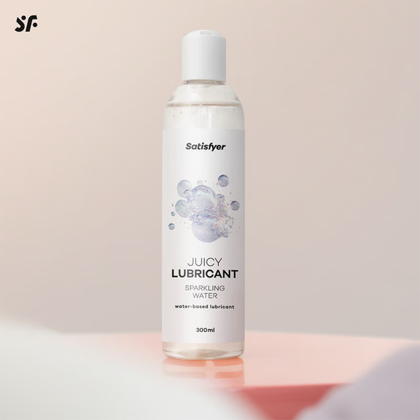 Studio product shot of the 300 ml Satisfyer Juicy Lubricant Sparkling Water bottle standing upright against a warm neutral background with a pink pedestal accent.