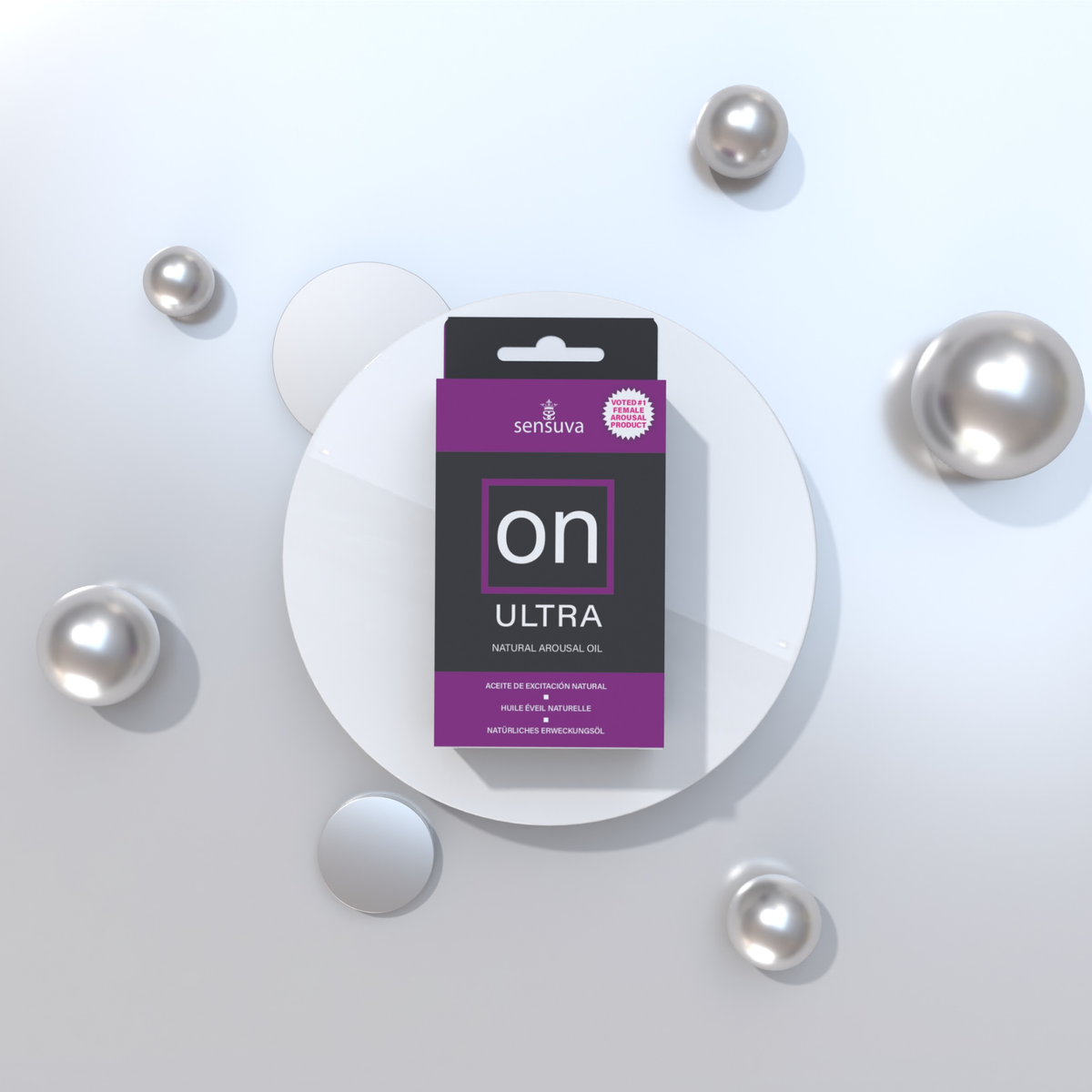 The packaged box of Sensuva ON Ultra Natural Arousal Oil sits centered on a round white pedestal. Reflective silver spheres of various sizes are scattered around the display against a clean, light grey background.