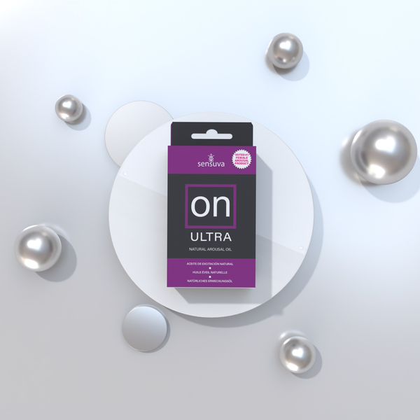 The packaged box of Sensuva ON Ultra Natural Arousal Oil sits centered on a round white pedestal. Reflective silver spheres of various sizes are scattered around the display against a clean, light grey background.