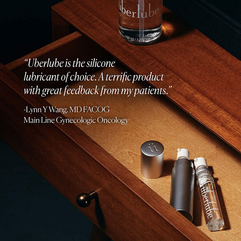 A clear bottle of "Uberlube" sits on a wooden drawer, with two other Uberlube products (one clear and one silver) partially visible inside the open drawer. A testimonial reads, "Uberlube is the silicone lubricant of choice. A terrific product with great feedback from my patients." -Lynn Y Wang, MD FACOG, Main Line Gynecologic Oncology, a product available at Worldcondoms.
