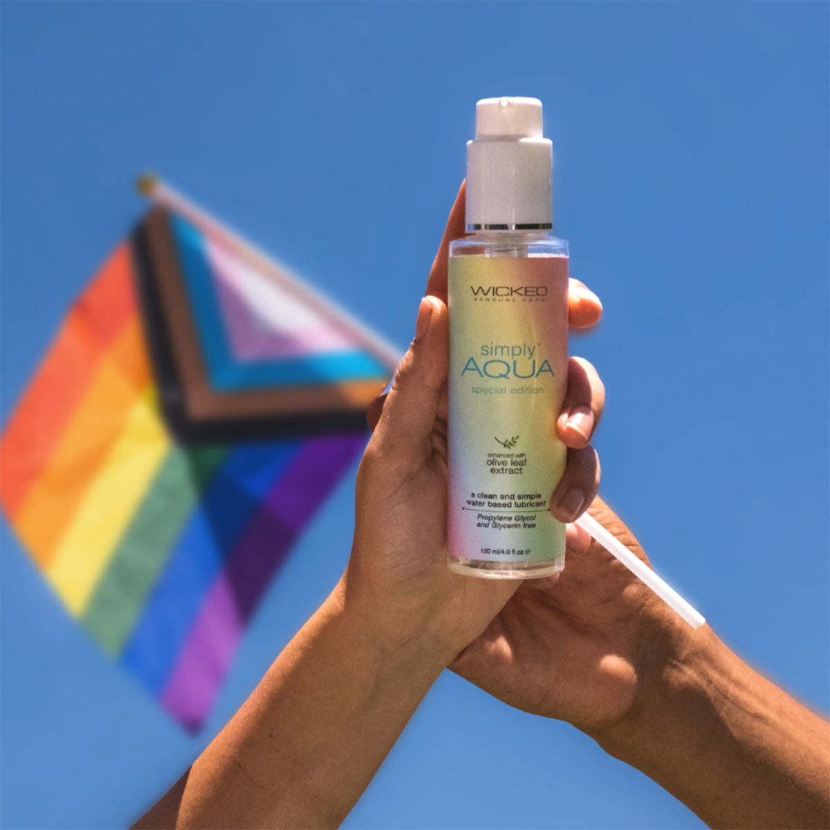 Two hands hold a bottle of Wicked Sensual Care "Simply Aqua" Special Edition lubricant upward against a clear blue sky. In the background, a Pride flag blurs in the wind. The bottle features a metallic rainbow-gradient label and states it is "enhanced with olive leaf extract" and "Propylene Glycol and Glycerin free."