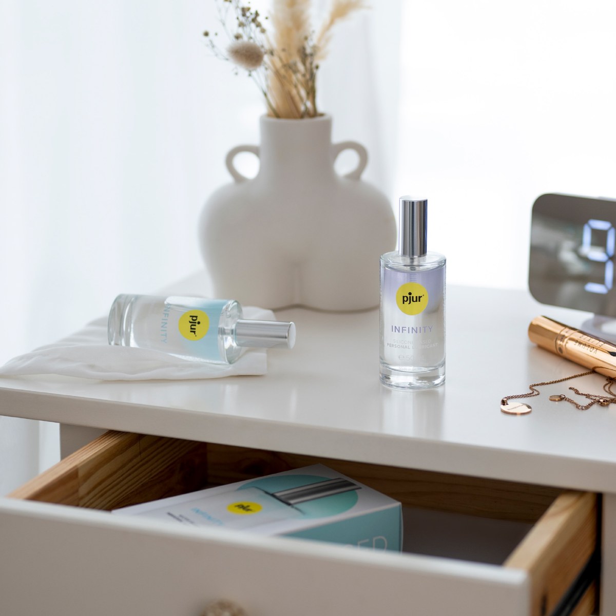 A lifestyle photo of two bottles of pjur Infinity Silicone-Based and Water-based Personal Lubricant sitting on a white nightstand. One bottle is standing upright, and the other is lying on its side on a white cloth. 