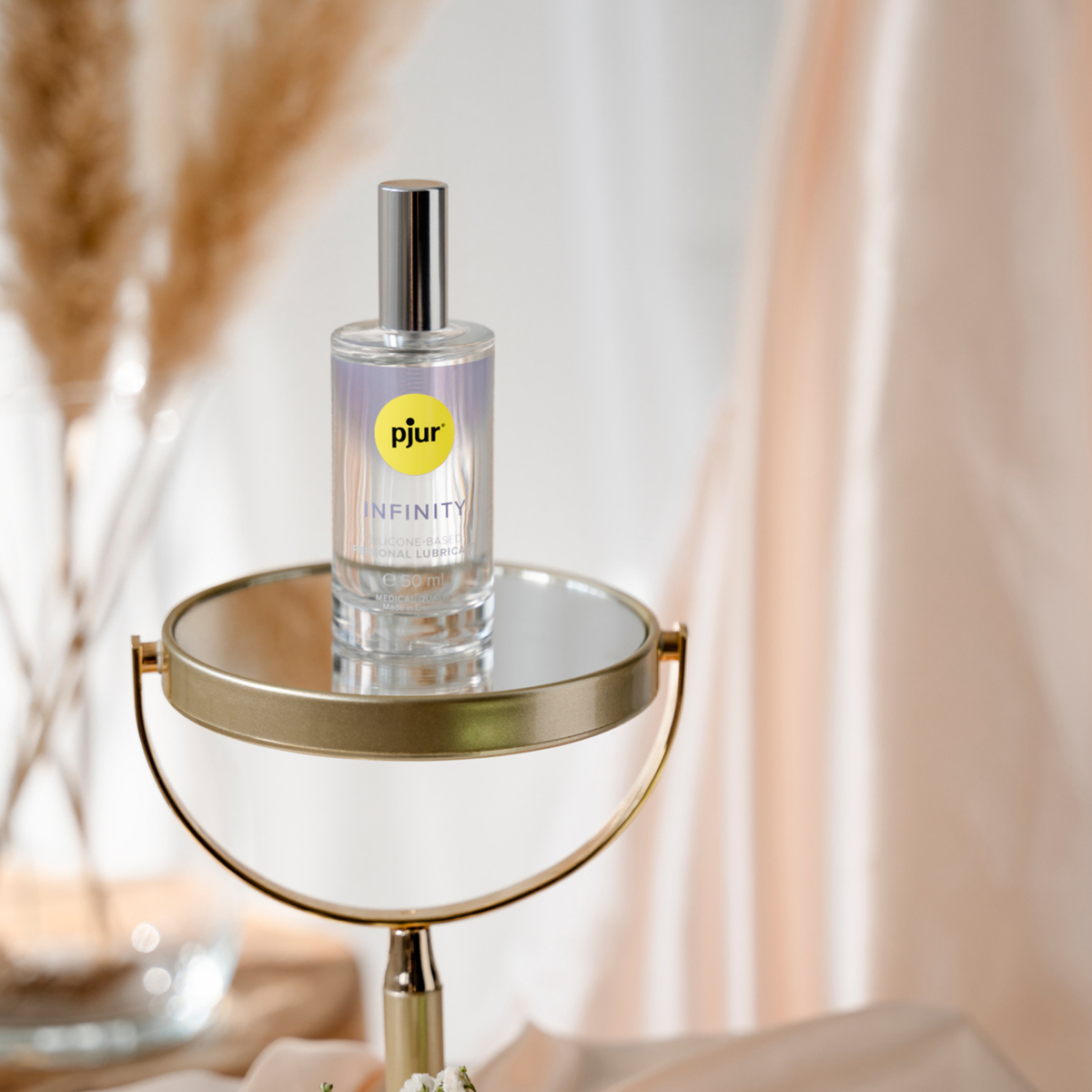 A clear bottle of Pjur Infinity lubricant with a silver pump rests on a small, round, mirrored table.
