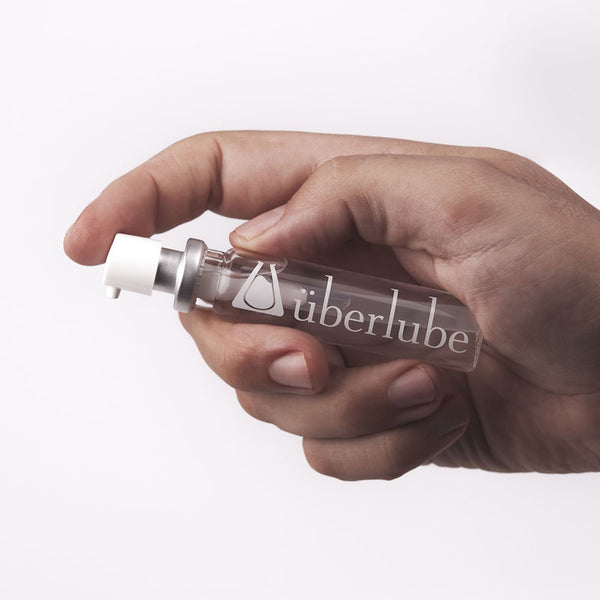 A hand holds a sleek, cylindrical dispenser with a white pump top, uberlube  personal lubricant from Worldcondoms.