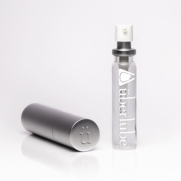 A tall, slender silver refillable case stands next to a clear glass bottle of "überlube" with a white pump top, both items for personal lubricant available at Worldcondoms.
